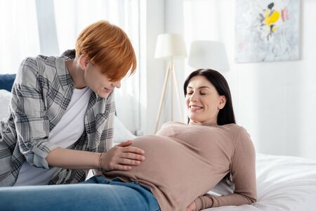 Smiling woman touching belly of pregnant girlfriend on bedの写真素材