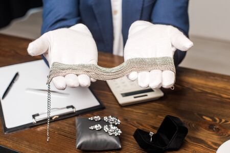 Cropped view of jewelry appraiser showing necklace near calculator, clipboard and jewelry on wooden tableの写真素材