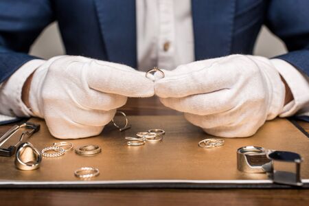 Cropped view of jewelry appraiser holding jewelry ring near board and magnifying glass on table isolated on greyの写真素材