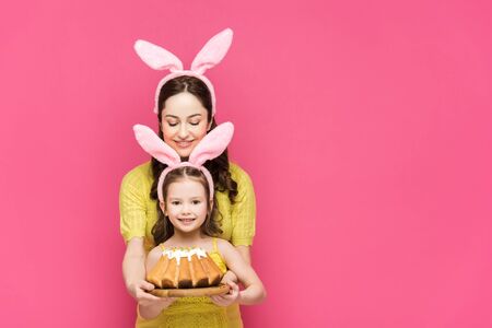 happy mother in bunny ears near daughter with easter cake isolated on pink