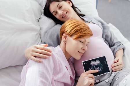 Smiling woman holding ultrasound scan of baby near belly of pregnant girlfriend on bed