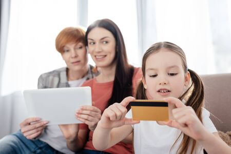 Selective focus of kid holding credit card near mothers using digital tablet on couchの写真素材