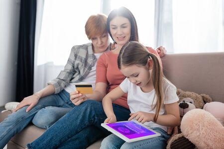 Child using digital tablet with shopping app on screen near same sex parents with credit card and soft toys on couchの写真素材