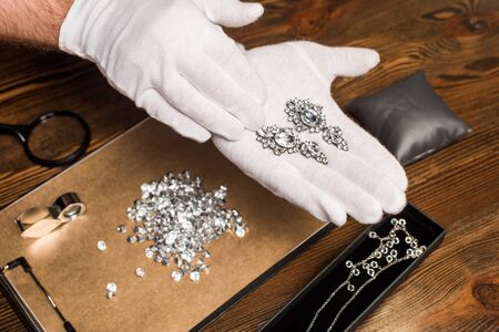 Cropped view of jewelry appraiser holding earrings near gemstones and magnifying glasses on board on...