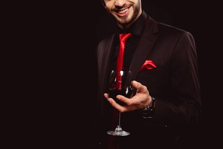 Cropped view of elegant man smiling while holding glass of red wine isolated on blackの写真素材
