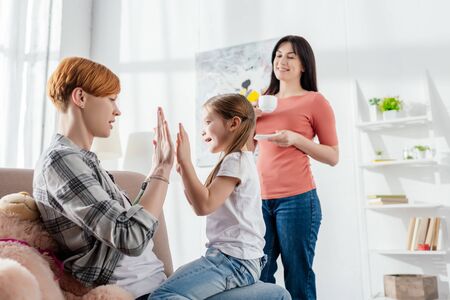 Mother playing with daughter on couch near smiling parent with coffee cup in living roomの写真素材