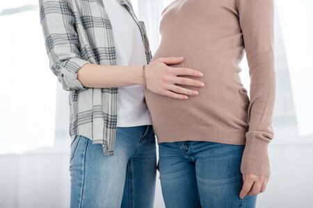 Cropped view of woman touching tummy of pregnant girlfriend at homeの写真素材