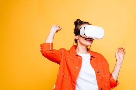 smiling girl gesturing while using vr headset isolated on yellow