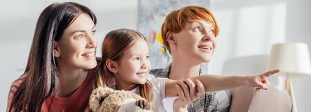 Panoramic shot of smiling kid with teddy bear pointing with finger near mothers on couch