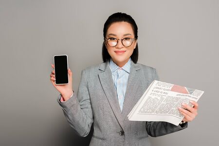 young businesswoman in eyeglasses holding newspaper and smartphone on grey background