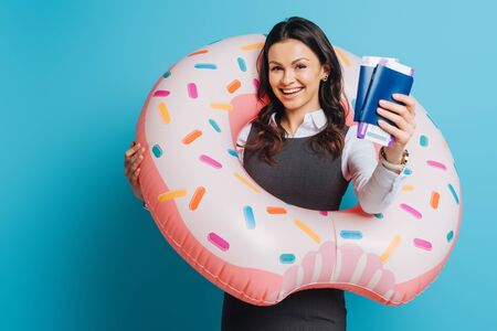 cheerful businesswoman in swim ring showing passports and air tickets on blue backgroundの写真素材