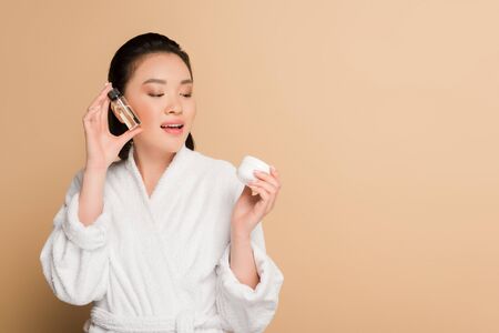 smiling beautiful asian woman in bathrobe holding cosmetic cream and oil on beige backgroundの写真素材