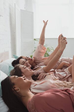Selective focus of multicultural women taking selfie and lying on bed at bachelorette partyの写真素材