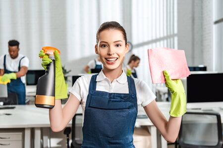 happy cleaner in overalls holding spray bottle and rag while looking at cameraの写真素材