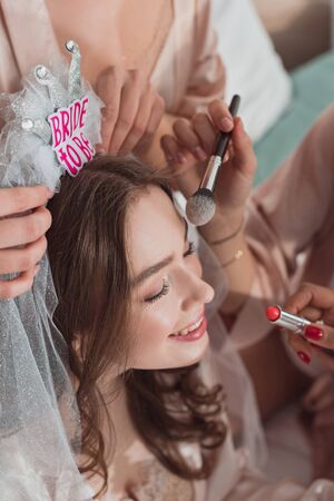 Cropped view of multicultural women putting makeup with cosmetic brush and lipstick on bride at bachelorette partyの写真素材