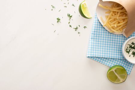top view of carton box with crispy french fries near garlic sauce and sliced lime on white
