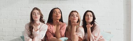 African american and caucasian women looking at camera and blowing kisses on bed at bachelorette party, panoramic shotの写真素材