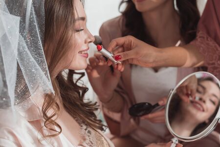 Cropped view of multiethnic women putting makeup with cosmetic brush and lipstick on bride on white backgroundの写真素材