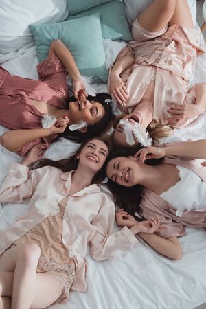 Top view of happy and excited multiethnic women resting on bed with feathers at bachelorette partyの写真素材