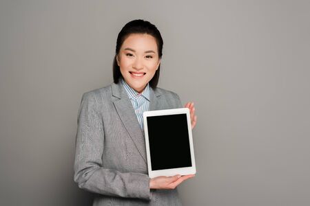 smiling young businesswoman in suit holding digital tablet on grey background