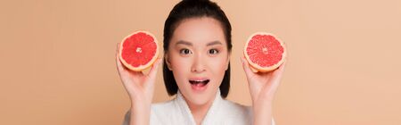 shocked beautiful asian woman in bathrobe with grapefruit halves isolated on beige, panoramic shotの写真素材