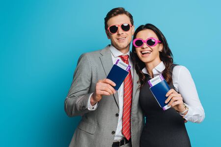 happy businessman and businesswoman in sunglasses holding passports and air tickets on blue backgroundの写真素材
