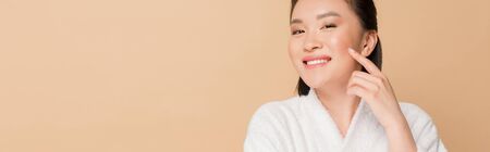 smiling beautiful asian woman in bathrobe touching face isolated on beige, panoramic shotの写真素材
