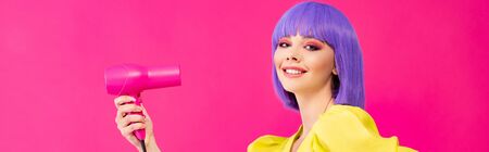 panoramic shot of cheerful pop art girl in purple wig using hair dryer, isolated on pinkの写真素材