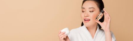 smiling beautiful asian woman in bathrobe holding cosmetic cream and oil isolated on beige, panoramic shotの写真素材