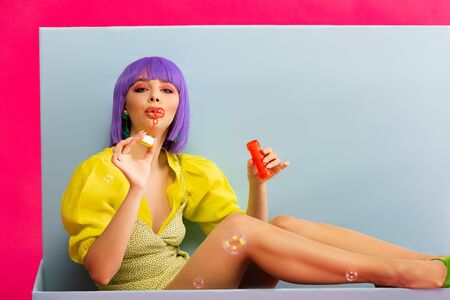 beautiful girl in purple wig as doll blowing soap bubbles while sitting in blue box, isolated on pinkの写真素材