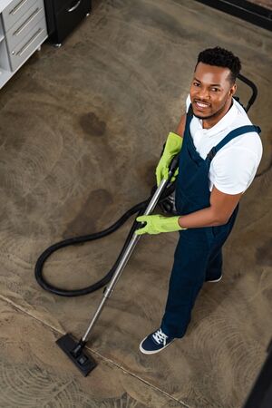 high angle view of smiling african american cleaner vacuuming floor while looking at cameraの写真素材