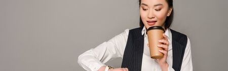 smiling young businesswoman looking at wristwatch and drinking coffee isolated on grey, panoramic shotの写真素材