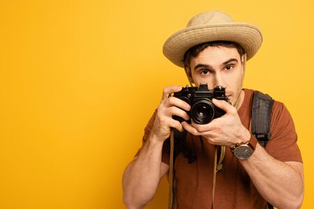 handsome tourist in hat with backpack taking photo on camera on yellow