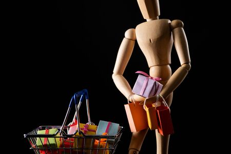 Wooden doll with shopping bags and presents in shopping basket isolated on blackの写真素材