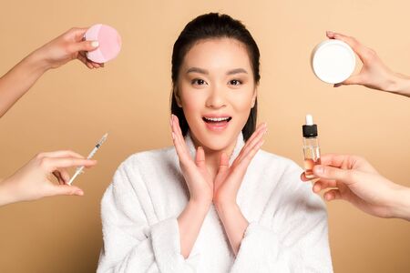 surprised beautiful asian woman in bathrobe near hands with facial cleansing brush, syringe, cosmetic cream and oil on beige backgroundの写真素材