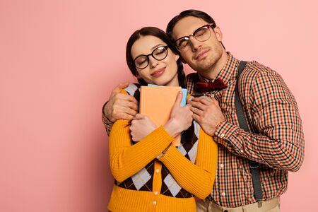 happy nerd with closed eyes hugging girlfriend with books on pink
