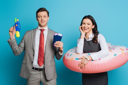 smiling businessman holding passports, air tickets and water gun near joyful businesswoman in swim ring on blue backgroundの写真素材