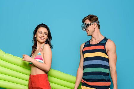 smiling man in diving mask looking at cheerful woman holding inflatable mattress isolated on blueの写真素材
