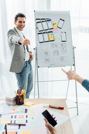Selective focus of smiling ux designer near whiteboard with templates pointing on colleague with...