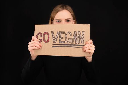 woman with obscure face holding cardboard with go vegan inscription isolated on black