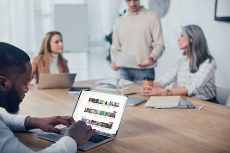 KYIV, UKRAINE - DECEMBER 6, 2019: selective focus of african american man using laptop with youtube website and his colleagues talking on backgroundのeditorial素材