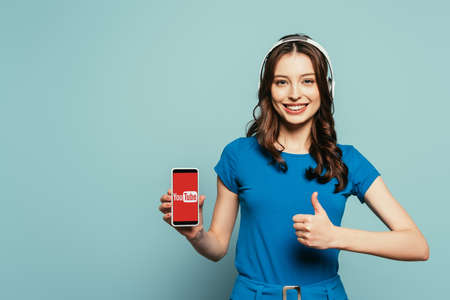 KYIV, UKRAINE - NOVEMBER 29, 2019: happy girl in wireless headphones showing thumb up while holding smartphone  with Youtube app on screen on blue backgroundのeditorial素材