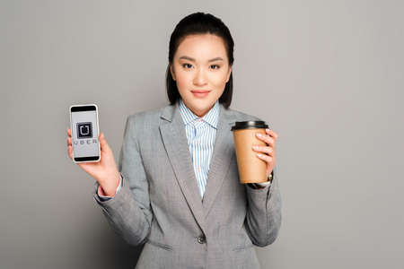 KYIV, UKRAINE -  FEBRUARY 11, 2019: happy young businesswoman with paper cup holding smartphone with uber app on grey backgroundのeditorial素材