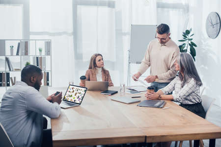 KYIV, UKRAINE - DECEMBER 6, 2019: colleagues doing paper work and african american man using smartphone near laptop with pinterest websiteのeditorial素材