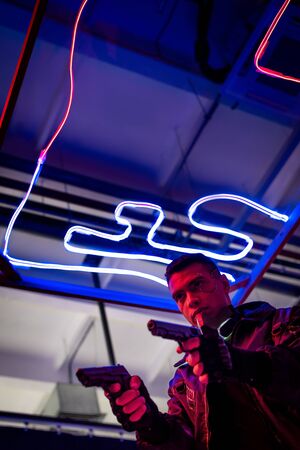 low angle view of armed bi-racial cyberpunk player holding guns near neon lightingの写真素材