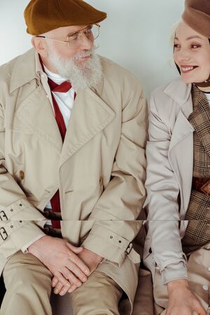 Stylish senior couple smiling at each other while sitting on white surface on grey background