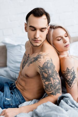 Side view of handsome tattooed man sitting back to back with attractive girl on bedの写真素材