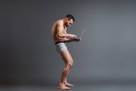 bearded and stooped man in boxer shorts using laptop on greyの写真素材