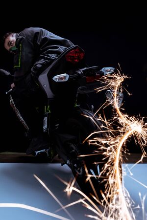 selective focus of mixed race cyberpunk player in mask riding motorcycle with sparks on black and...