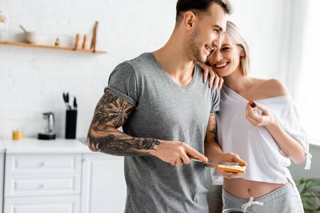 Beautiful smiling woman holding grape and embracing tattooed boyfriend with toast in kitchenの写真素材